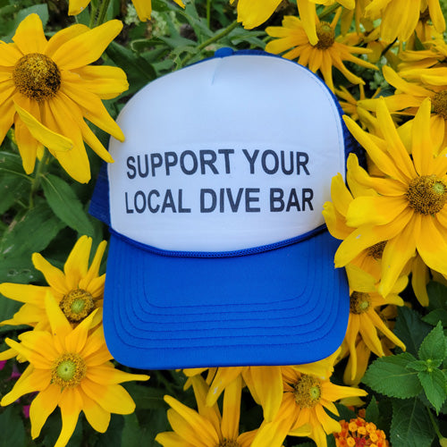 Blue trucker hat with text "support your local dive bar" on front of hat, sitting in sunflower