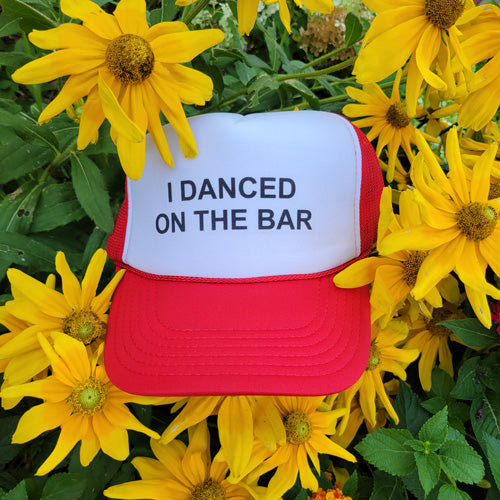 Red trucker hat with text "i danced on the bar" on front of hat, sitting in sunflowers
